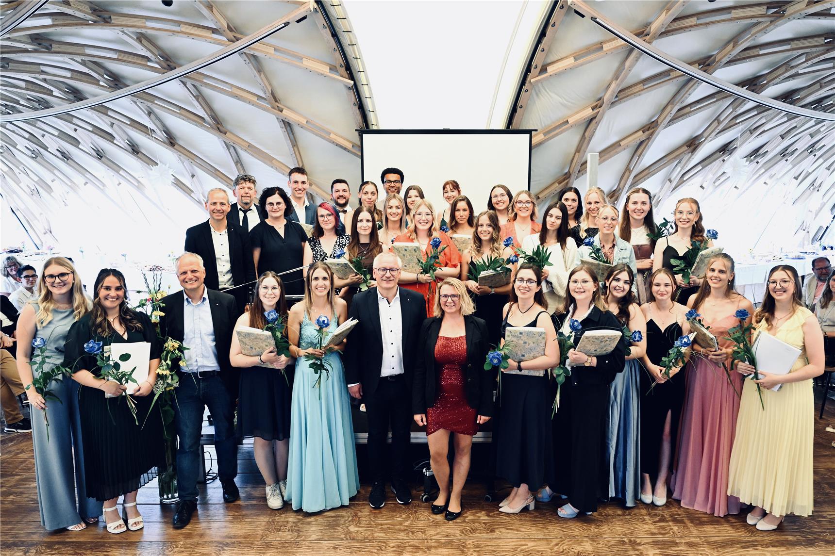 Eine große Gruppe von Menschen, zumeist Frauen in formellen Kleidern, posiert in einem Innenraum mit Blumen und Urkunden. Ein paar Männer in Anzügen stehen zwischen ihnen. Sie lächeln unter einer gewölbten Holzdecke.