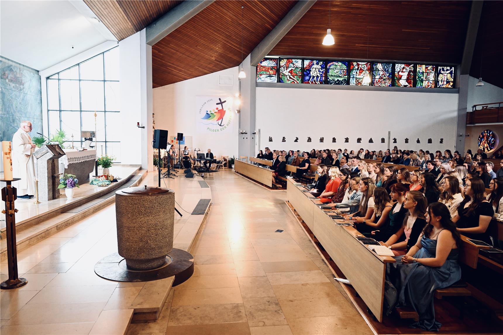 Eine Gemeinde sitzt in den Kirchenbänken einer modernen Kirche mit hohen Fenstern und bunten Glasfenstern. Ein Priester steht am Altar und spricht, während die Menschen aufmerksam zuhören. Der Innenraum ist geräumig und hat eine hohe Holzdecke.