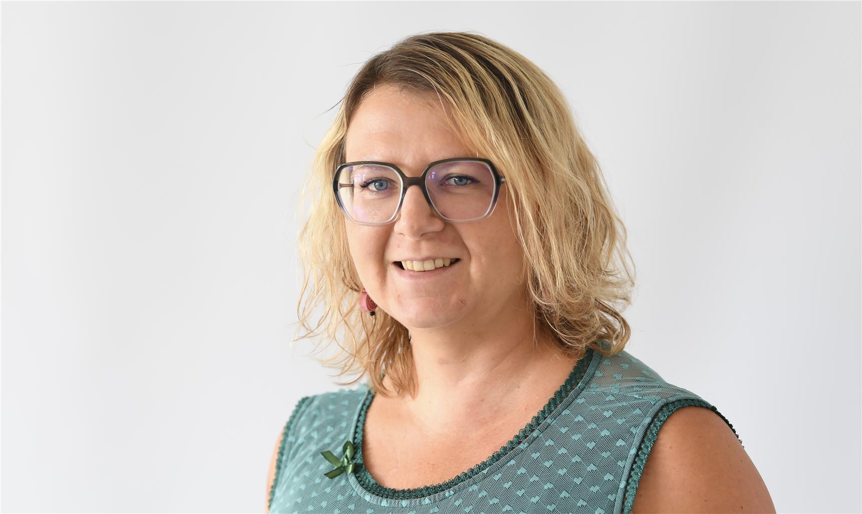 Eine Frau mit schulterlangen blonden Haaren, die eine Brille und ein ärmelloses grün gemustertes Oberteil trägt, lächelt in die Kamera vor einem schlichten weißen Hintergrund.