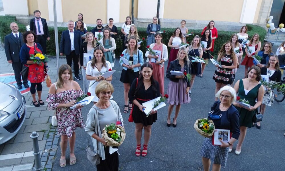 Eine Gruppe von Menschen, überwiegend Frauen in formeller Kleidung, steht draußen auf dem Bürgersteig, hält Blumen und Urkunden in den Händen und lächelt in die Kamera. Wahrscheinlich feiern sie eine Abschluss- oder Preisverleihung.