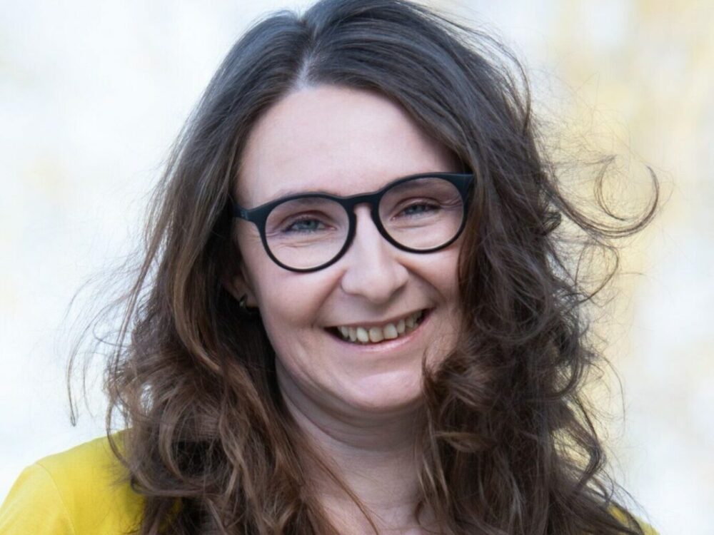 Eine Frau mit langen, welligen braunen Haaren und schwarzer Brille lächelt in die Kamera. Sie trägt ein gelbes Oberteil und ist vor einem leicht verschwommenen hellen Hintergrund abgebildet.