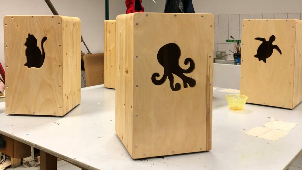Auf einem Tisch stehen drei hölzerne Cajón-Trommeln. Jede ist mit einer anderen Tiersilhouette versehen: einer Katze, einem Oktopus und einer Schildkröte. Im Hintergrund sind eine geflieste Wand und Werkstattmaterialien zu sehen.