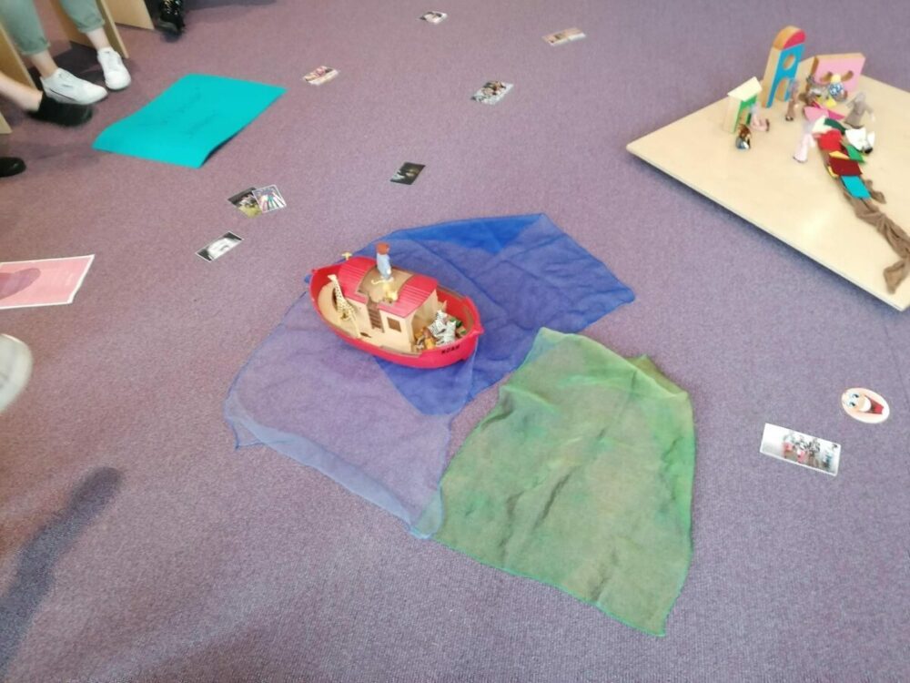 Eine Spielzeugarche mit Tierfiguren steht auf blauen und grünen Tüchern, die an Wasser und Land erinnern, auf einem Teppichboden. In der Nähe sind Fotos verstreut, und am Rand der Szene liegen Füße von Menschen.