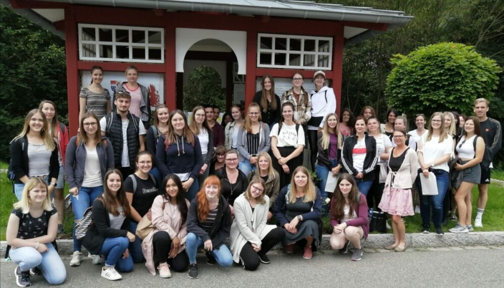 Eine große Gruppe von Menschen, hauptsächlich junge Frauen, posiert draußen vor einem rot-weißen Gebäude, umgeben von Bäumen und Grün. Sie lächeln und scheinen an einem bewölkten Tag ein Gruppenfoto zu machen.