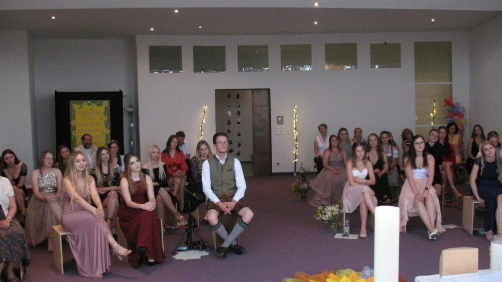 Eine Gruppe von Menschen, hauptsächlich junge Frauen in Abendkleidern und ein Mann in traditioneller Kleidung, sitzen in einem dekorierten Raum mit violettem Teppich, Blumen und Lichterketten und blicken in die Kamera, als würden sie einer Zeremonie oder Veranstaltung beiwohnen.