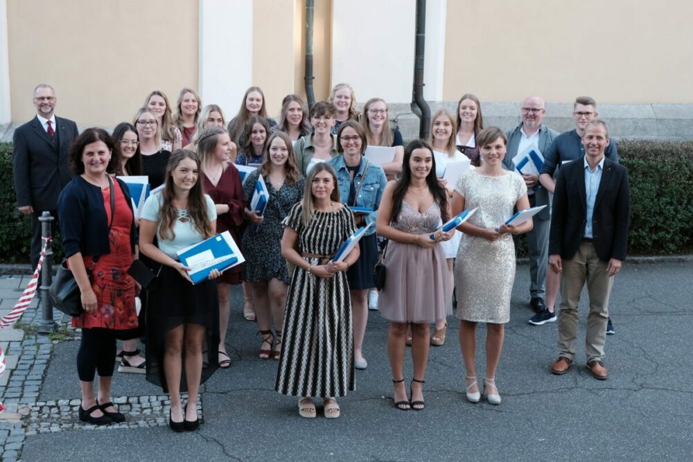 Eine Gruppe von Menschen, hauptsächlich Frauen, steht draußen vor einem Gebäude und lächelt in die Kamera. Einige halten blaue Ordner in den Händen, was auf eine Abschlussfeier oder Preisverleihung schließen lässt. Die Atmosphäre wirkt formell, aber freundlich.