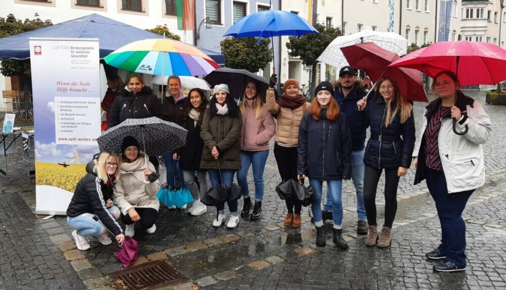 Eine Gruppe von Menschen steht unter bunten Regenschirmen auf einer nassen Kopfsteinpflasterstraße und lächelt vor einem Stand mit einem großen Banner in die Kamera. Einige tragen Jacken und Hüte, was auf kühles, regnerisches Wetter hindeutet.