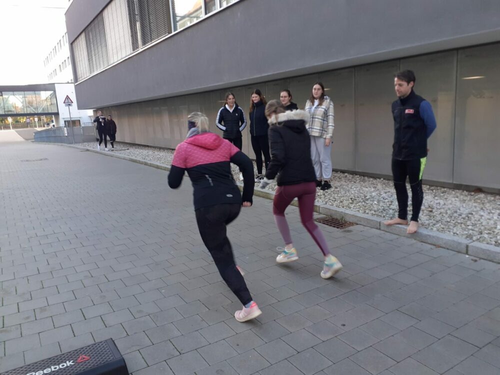 Zwei Frauen in Sportkleidung laufen im Freien auf einer gepflasterten Fläche, während eine Gruppe von sechs Personen, einige barfuß, in der Nähe vor einem modernen Gebäude steht und zusieht.