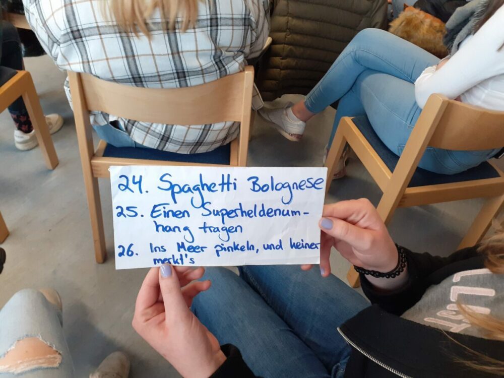 Eine Person hält eine handschriftliche Liste auf Deutsch mit den Nummern 24 bis 26 der Punkte. Im Hintergrund sitzen weitere Personen auf Stühlen und die Szene scheint in einem Klassenzimmer oder einer lockeren Gruppensituation zu spielen.