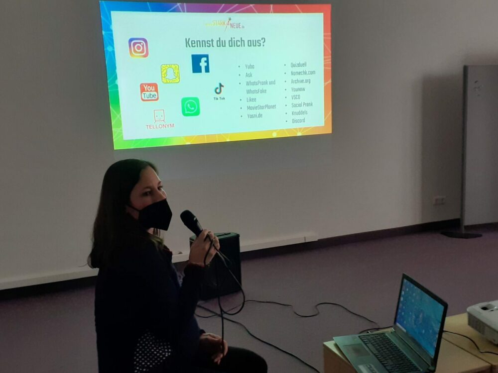 Eine Person mit Maske hält ein Mikrofon, sitzt neben einem Laptop und präsentiert eine Folie über Social-Media-Plattformen, die auf eine Wand in einem Klassenzimmer oder Besprechungsraum projiziert wird.
