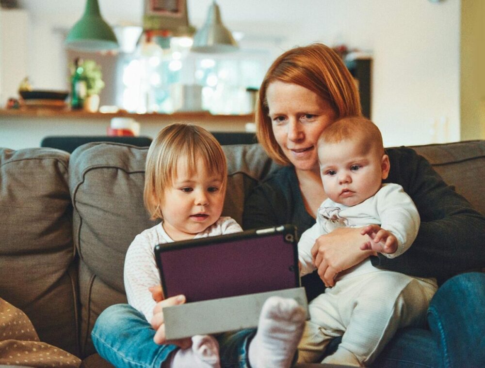 Eine Frau sitzt mit einem Baby im Arm und einem Kleinkind neben einem Sofa. Alle schauen gemeinsam in einem gemütlichen, gut beleuchteten Wohnzimmer auf ein Tablet.