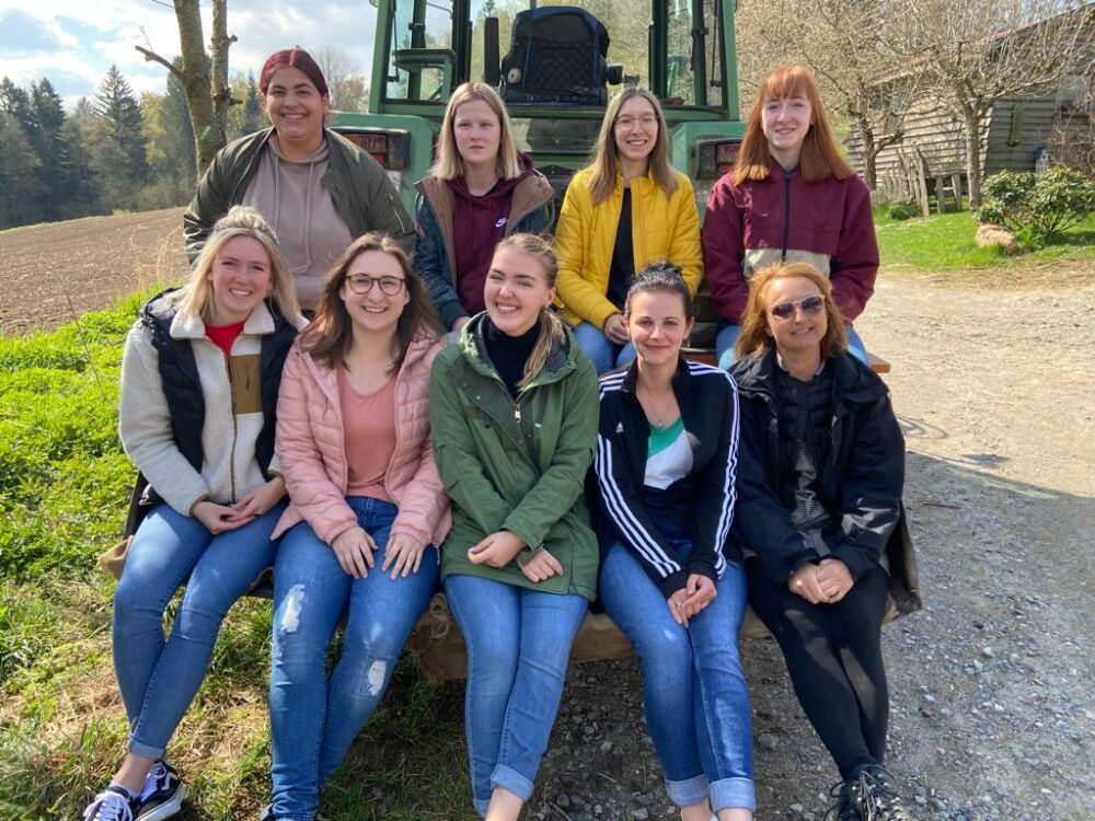 Neun Frauen lächeln und posieren an einem sonnigen Tag im Freien für ein Gruppenfoto. Sie sitzen und stehen vor einem grünen Traktor, im Hintergrund sind Bäume, ein Feld und ein rustikales Gebäude zu sehen.