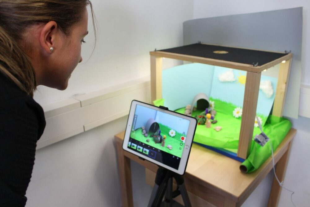 Eine Frau beobachtet ein Tablet auf einem Stativ, das eine Stop-Motion-Animationsszene mit Tonfiguren und einem bunten Hintergrund in einer kleinen Box auf einem Holztisch filmt.