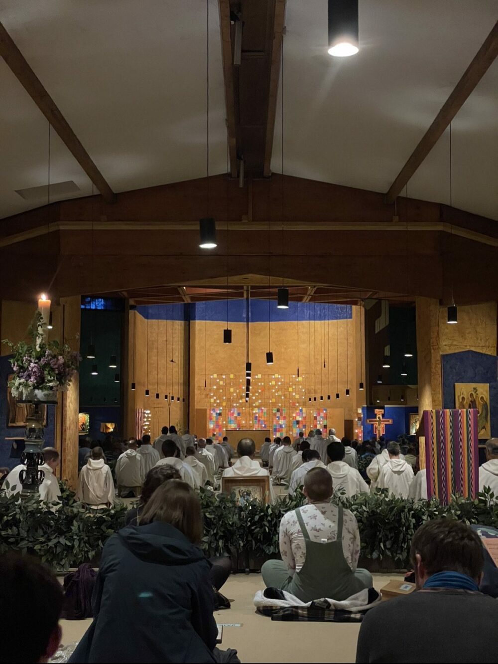 In einer Holzkirche sitzt eine Gemeinde vor einem mit bunten Dekorationen und Kerzen geschmückten Altar. Geistliche in weißen Gewändern sitzen vorne, Grünpflanzen und Blumen schmücken den Raum.
