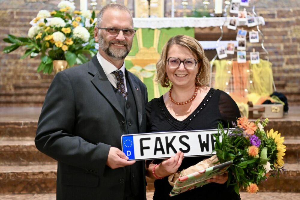 Ein Mann im Anzug und eine Frau mit Blumen lächeln und halten ein neuartiges deutsches Nummernschild mit der Aufschrift „FAKS WR“ in den Händen. Sie stehen vor einem mit Blumen und Fotos geschmückten Altar.