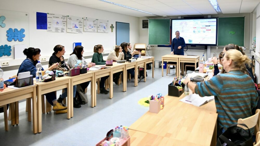 Eine Gruppe Erwachsener sitzt in einem Klassenzimmer an U-förmig angeordneten Tischen und hört einem Lehrer zu, der neben einem großen digitalen Bildschirm mit bunten Diagrammen und Texten steht. Auf den Tischen sind Unterrichtsmaterialien zu sehen.
