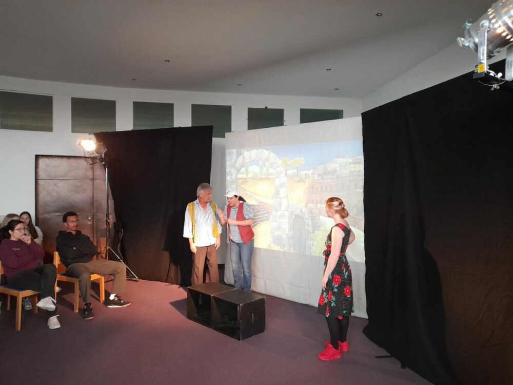 Drei Schauspieler führen ein Theaterstück auf einer kleinen Bühne mit projiziertem Hintergrund auf, während links ein kleines Publikum zusieht. Studiolichter und schwarze Vorhänge rahmen die Szene ein.