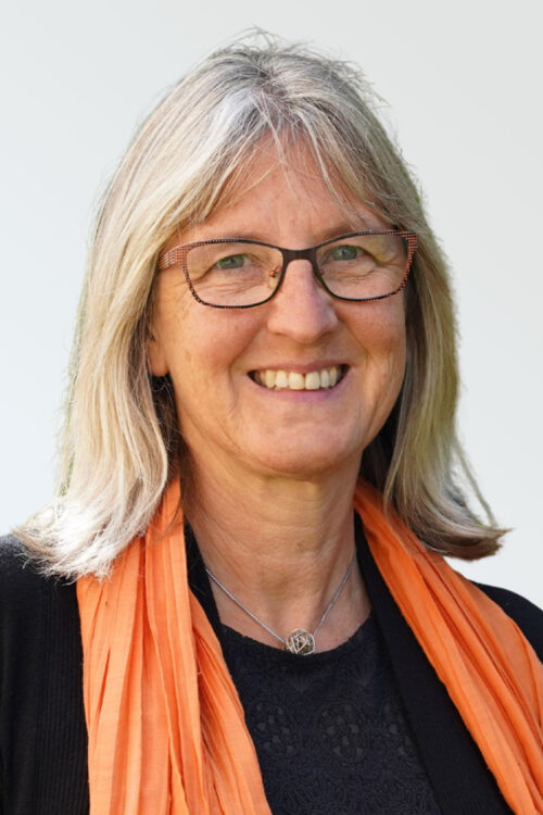 Eine lächelnde Frau mit schulterlangem grauem Haar, die eine Brille, ein schwarzes Oberteil und einen orangefarbenen Schal trägt, vor einem hellen Hintergrund.