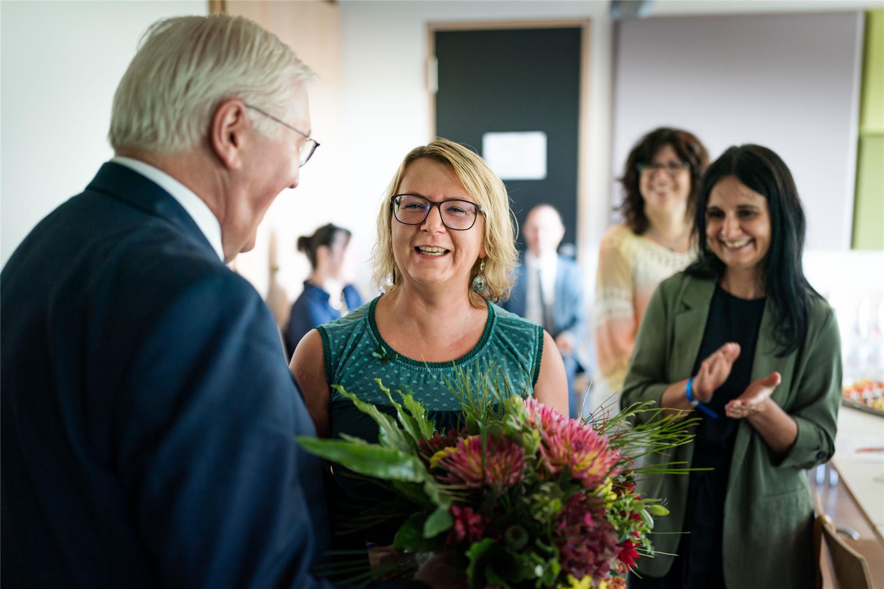 Eine Frau, die einen Blumenstrauß in der Hand hält, lächelt einen älteren Mann im Anzug an. Zwei Frauen im Hintergrund klatschen und lächeln in einem hellen Raum, was auf einen feierlichen Anlass hindeutet.